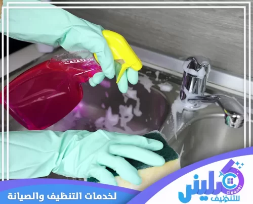 شخص متخصص في تنظيف المطابخ بجدة يرتدي قفازات زرقاء يرش سائل تنظيف على حوض، مع إسفنجة، كلينر للتنظيف لن تجد لها منافس 0500892799.