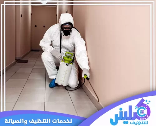 فني مكافحة حشرات من شركة كلينر بالمدينة المنورة أثناء رش المبيدات في زاوية ممر داخلي، مرتديًا زي الحماية الكامل لضمان السلامة. يركز على حواف الجدران والأرضيات باستخدام مرشة يدوية للقضاء على الحشرات الزاحفة مثل الصراصير والنمل، مما يعكس التزام الشركة بمعايير السلامة وتقنيات الرش الاحترافي.