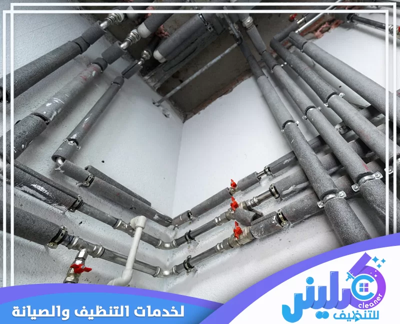 عزل المواسير بالصوف الصخري بالرياض 0500892799 شركة كلينر. شبكة مواسير مياه معزولة بغطاء رمادي مثبتة على حائط أبيض، مع ظهور محابس حمراء ومشابك معدنية.