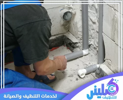 شركة عزل مواسير المياه بالرياض 0500892799 شركة كلينر. فني يركب عازل رمادي حول مواسير مياه داخل زاوية حائط مكسور، مع ظهور أكواع بيضاء.