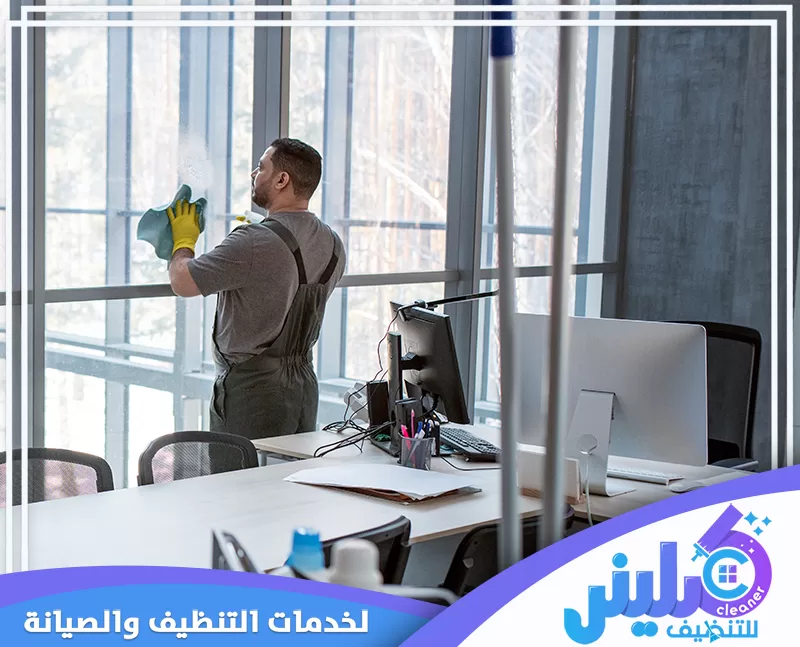عامل ينظف زجاج نافذة داخل مكتب حديث باستخدام قطعة قماش، وسط مكاتب وأجهزة كمبيوتر.