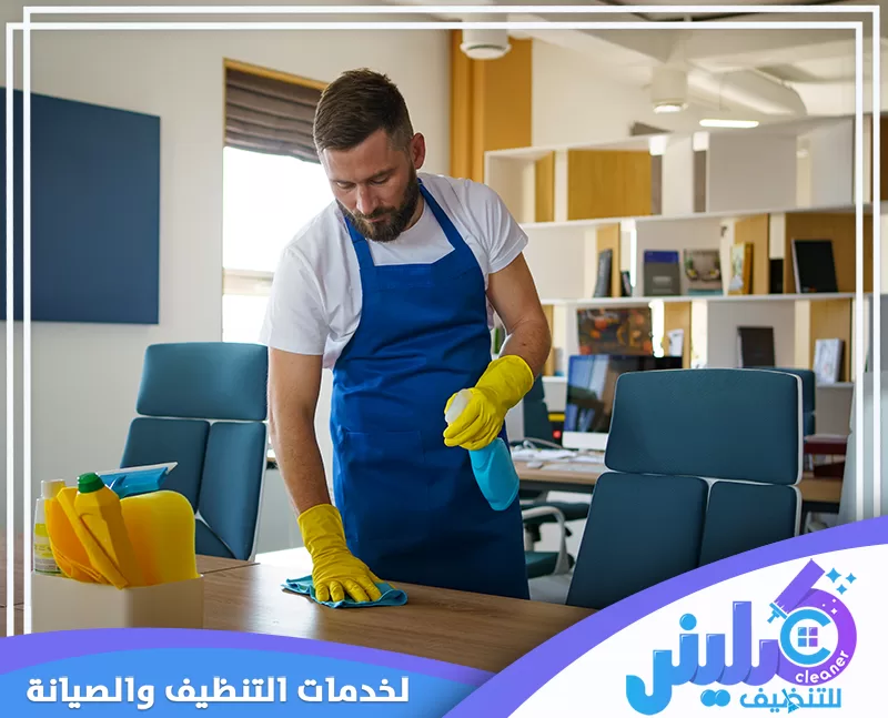عامل تنظيف مكاتب يمسح سطح المكتب ويستخدم بخاخ تنظيف داخل مساحة عمل حديثة ومنظمة.